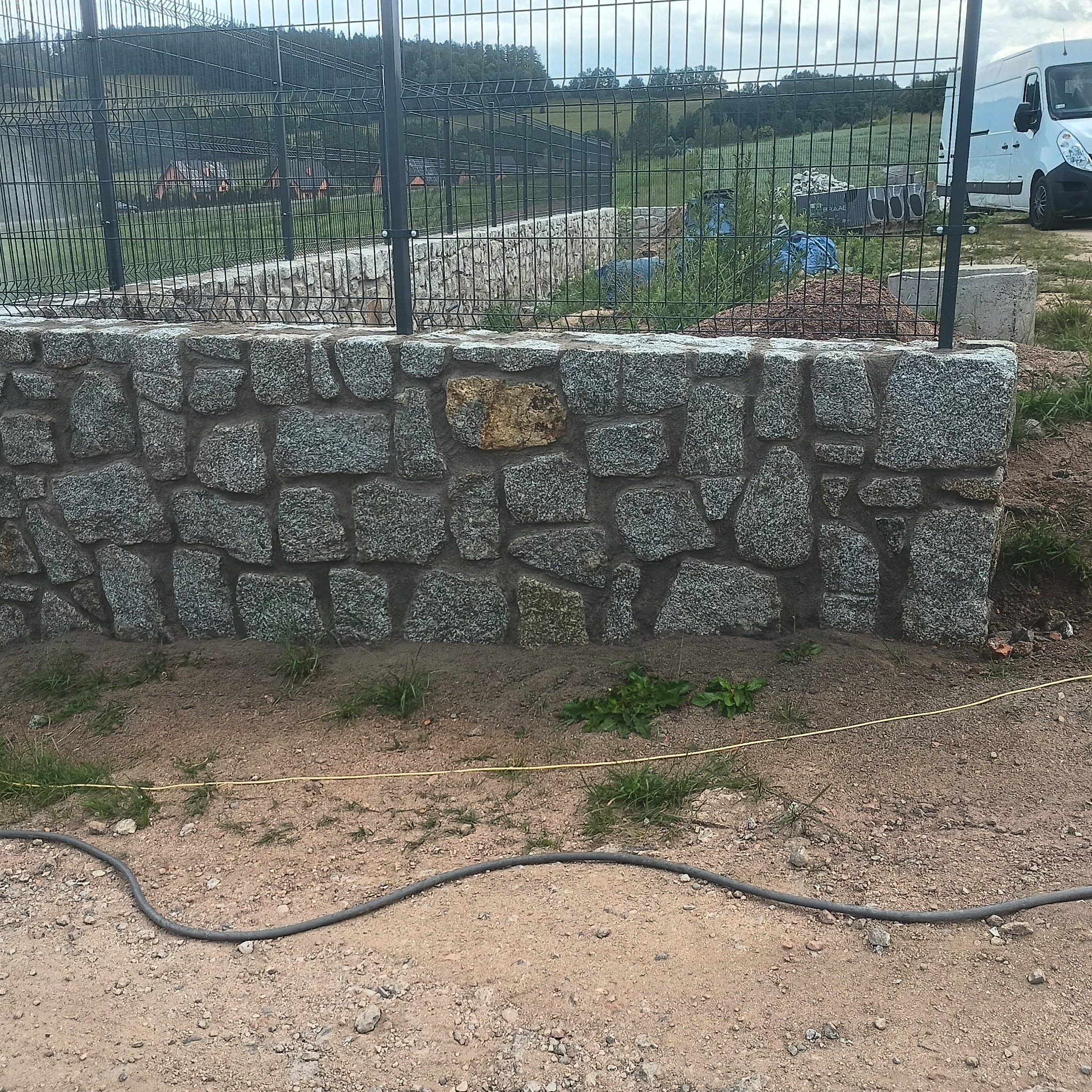 Murek oporowy z nieregularnych kamieni granitowych, z widoczną siatką ogrodzeniową w tle i kablem na ziemi.