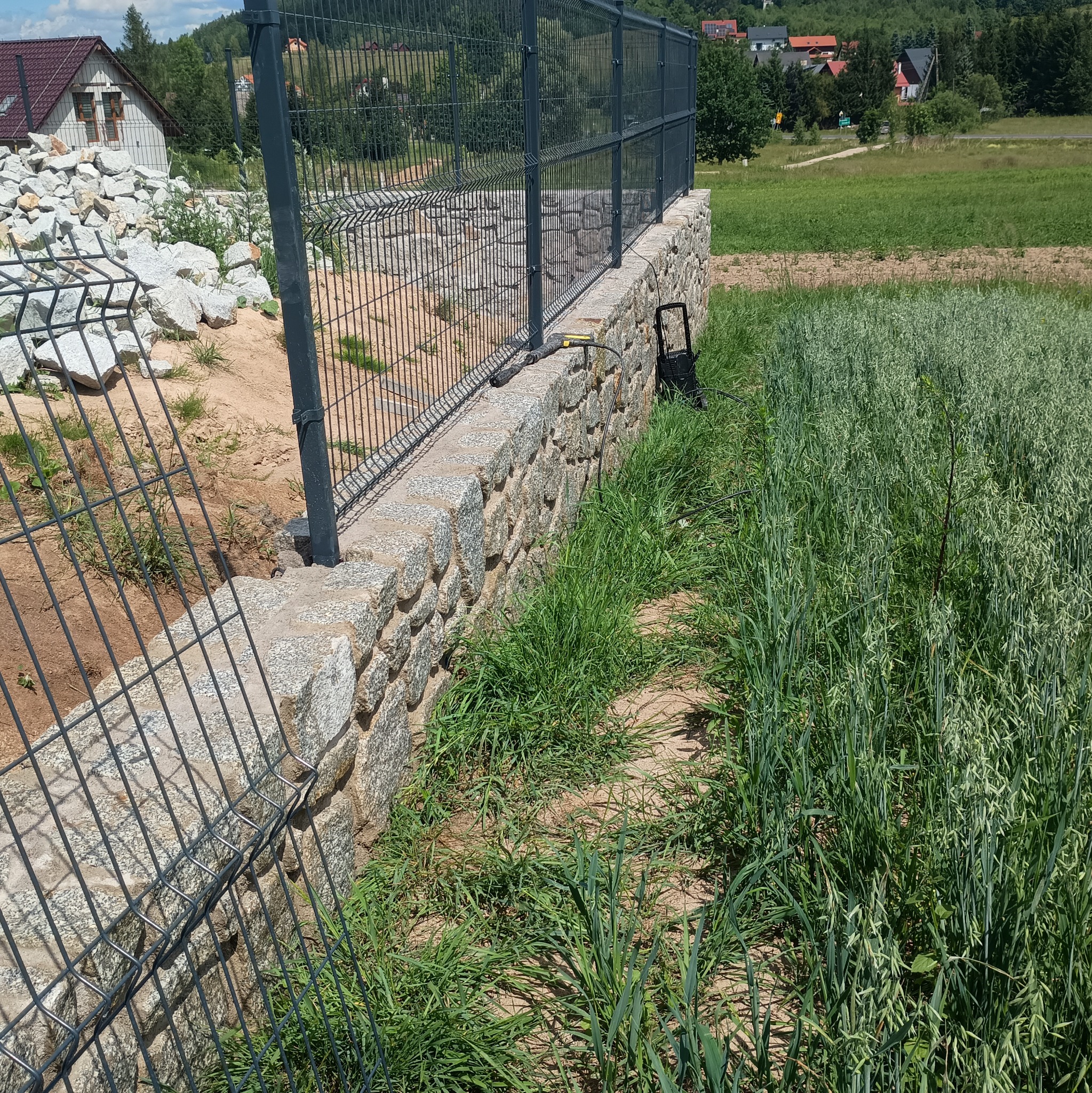 Metalowe ogrodzenie panelowe na kamiennej podmurówce z granitu, widoczna myjka ciśnieniowa i wysoka trawa w tle.