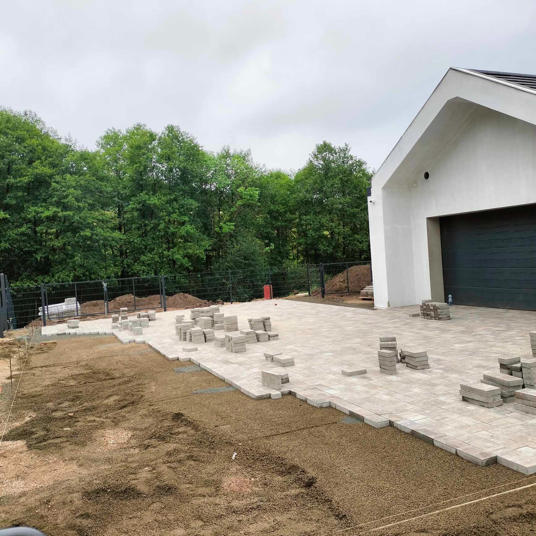 W Sosnówce trwa układanie beżowej kostki brukowej na przygotowanym podłożu. Część kostki ułożona, reszta w stosach. W tle nowoczesny dom z garażem i zieleń drzew.