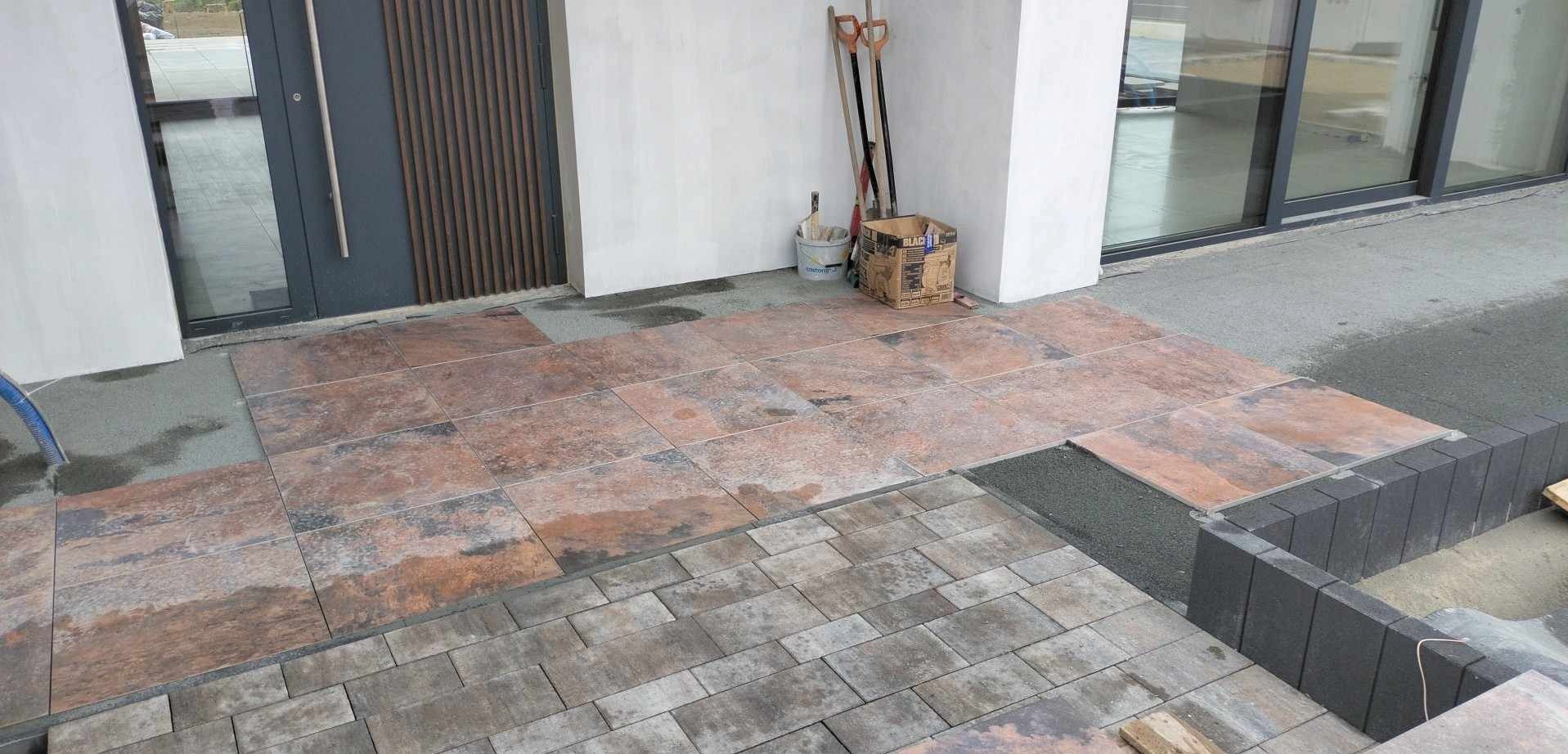 Fragment układania tarasu z płytek imitujących kamień, z widocznymi narzędziami i materiałami budowlanymi, w tle drzwi wejściowe i okno.