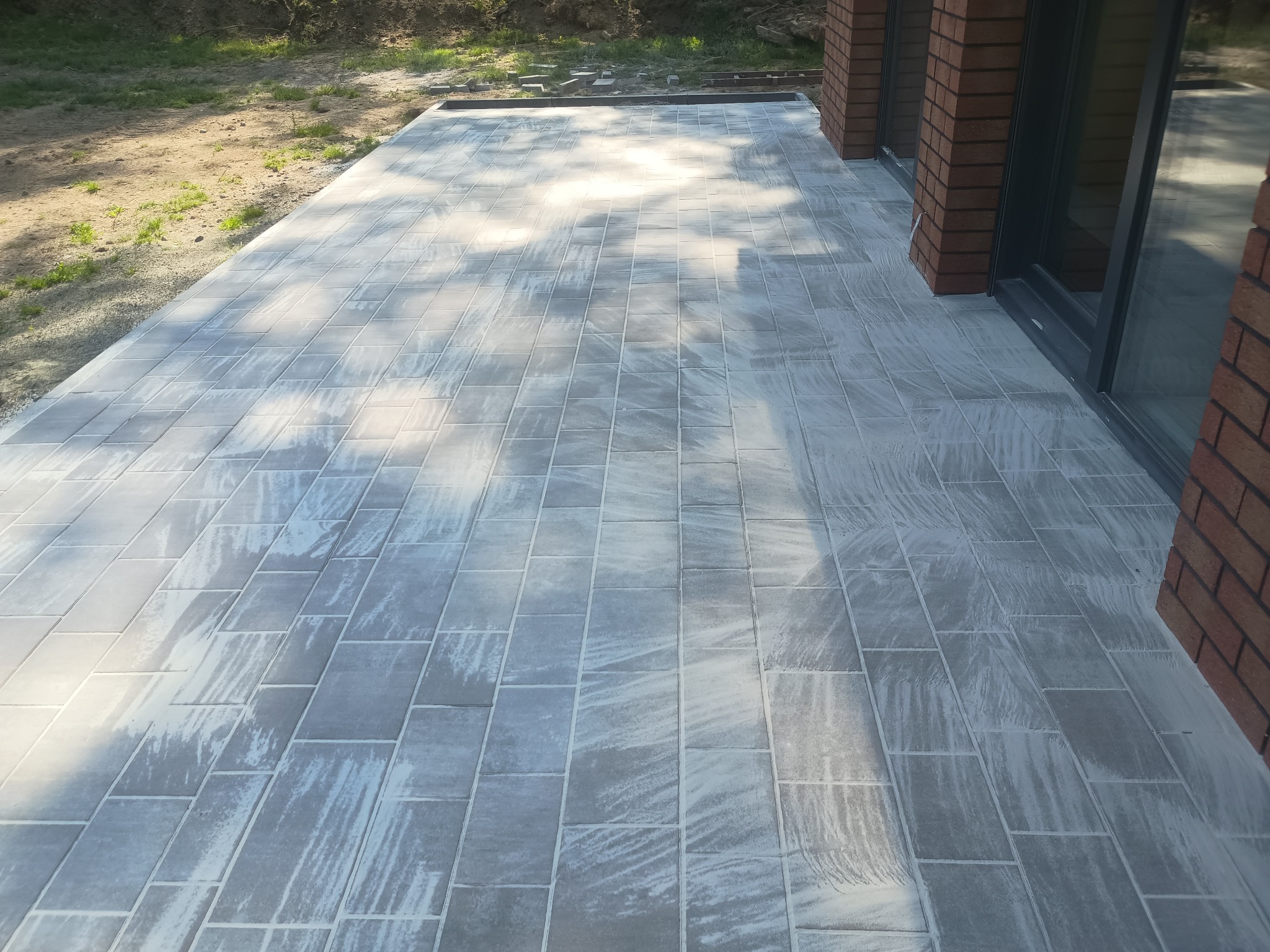 Świeżo ułożona, szara kostka brukowa w kształcie prostokątów, tworząca taras przylegający do budynku z ceglanymi ścianami i dużymi oknami.