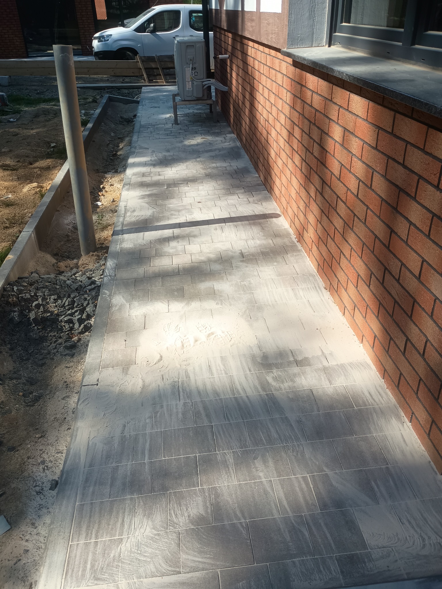 Ułożona szara kostka brukowa przy ścianie z czerwonej cegły, widoczny rów z kamieniami i zaparkowany biały samochód dostawczy w tle.
