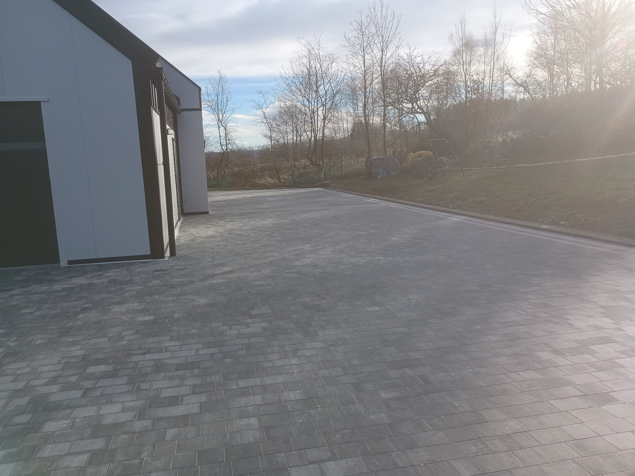 Ułożona szara kostka brukowa przed budynkiem w Sosnówce, widoczne drzewa w tle i promienie słońca.