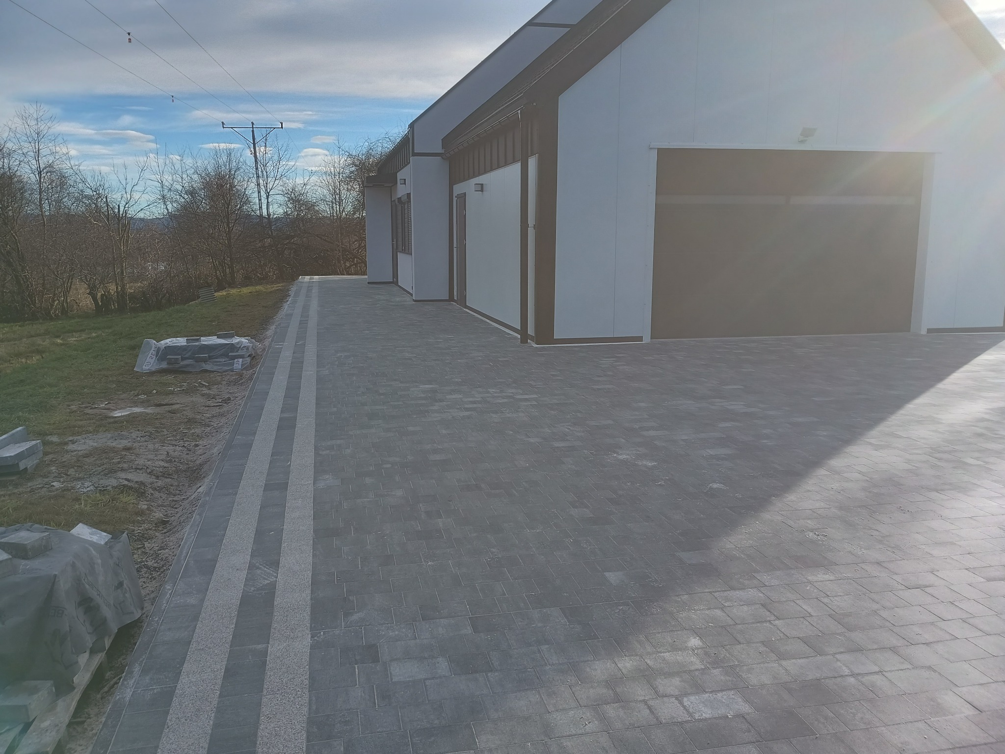 Ułożona szara kostka brukowa z jasnymi pasami dekoracyjnymi prowadząca do nowoczesnego budynku z bramą garażową, w tle drzewa i pochmurne niebo.