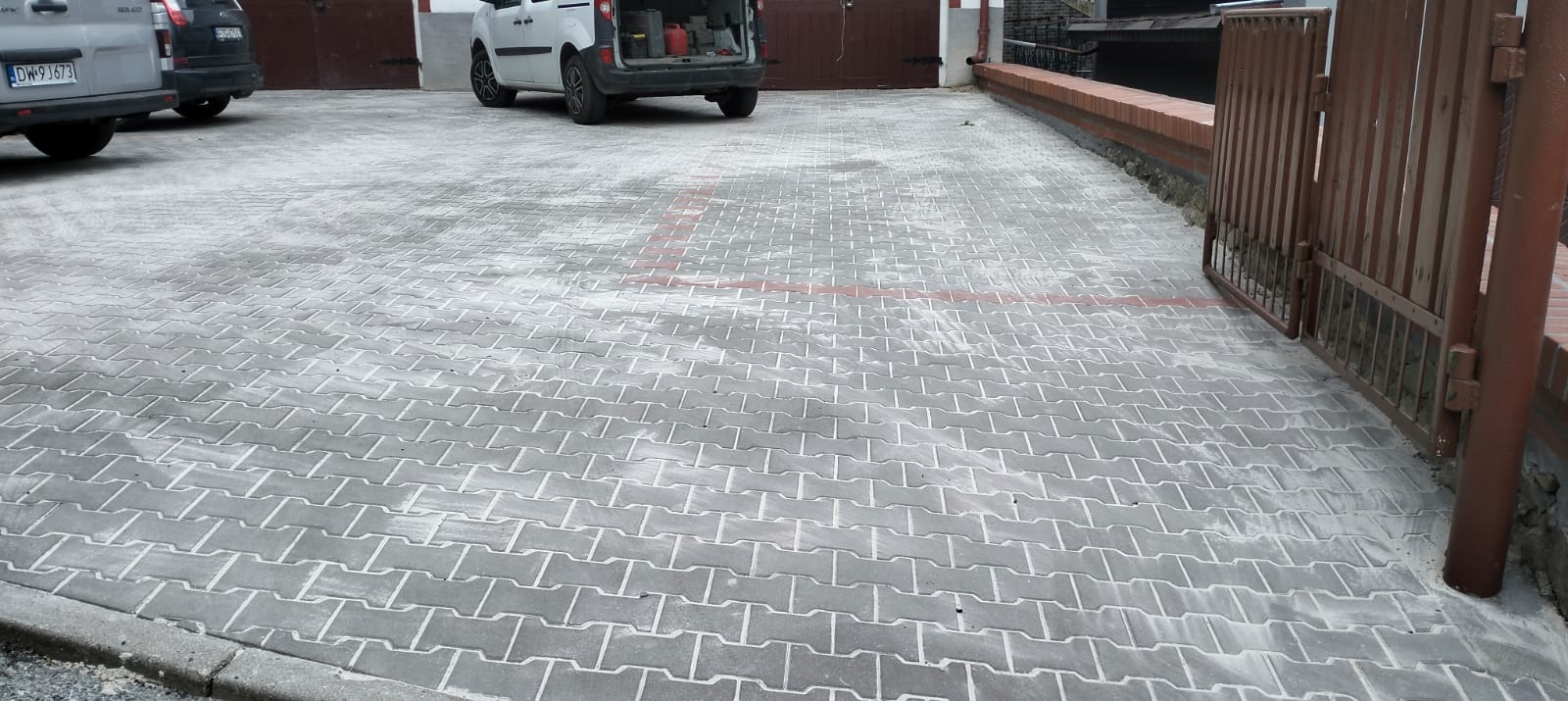 Szara kostka brukowa ułożona na podjeździe z widocznymi liniami parkingowymi, obok metalowa brama i zaparkowane samochody dostawcze.