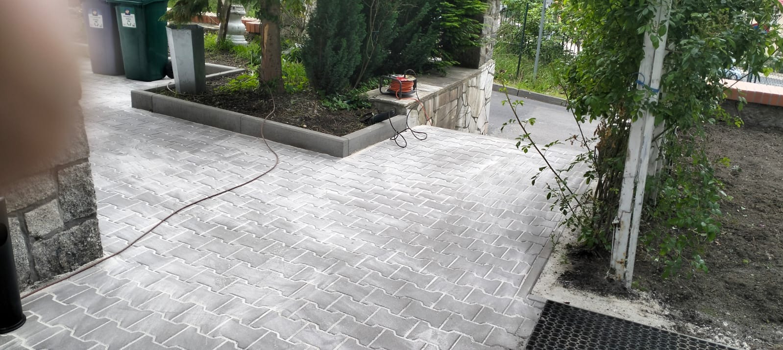 Ułożona szara kostka brukowa na posesji, obrzeża wykończone kostką, widoczne rośliny i elementy ogrodu. Na murku leży zwinięty przedłużacz.