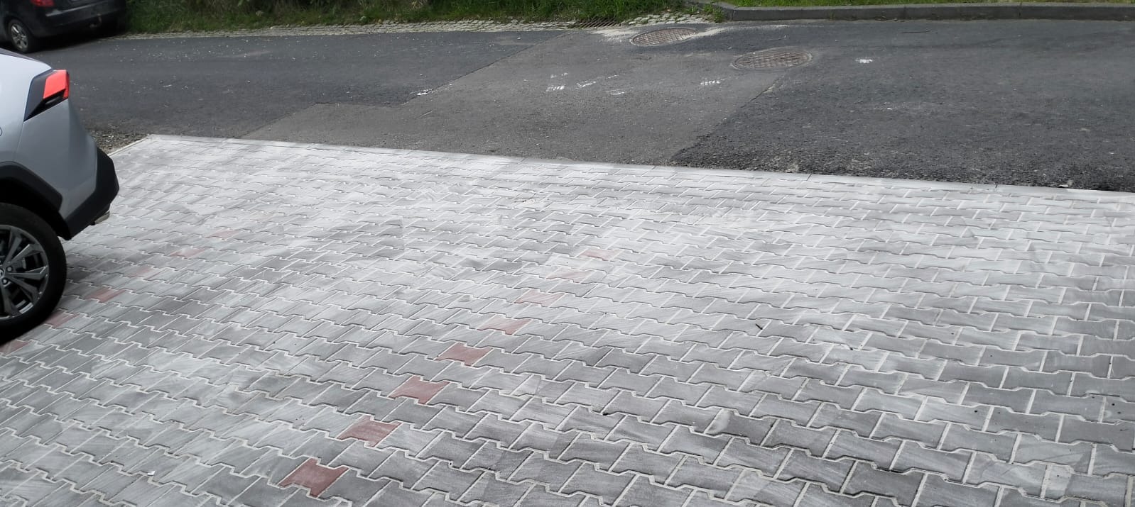 Ułożona szara kostka brukowa typu fala z czerwonymi akcentami, obok zaparkowany samochód. Widoczny fragment asfaltowej drogi.