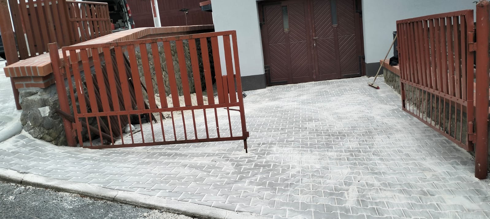 Ułożona szara kostka brukowa przed budynkiem z brązową bramą garażową. Widoczne elementy ogrodzenia z metalowymi furtkami w kolorze rdzawym.
