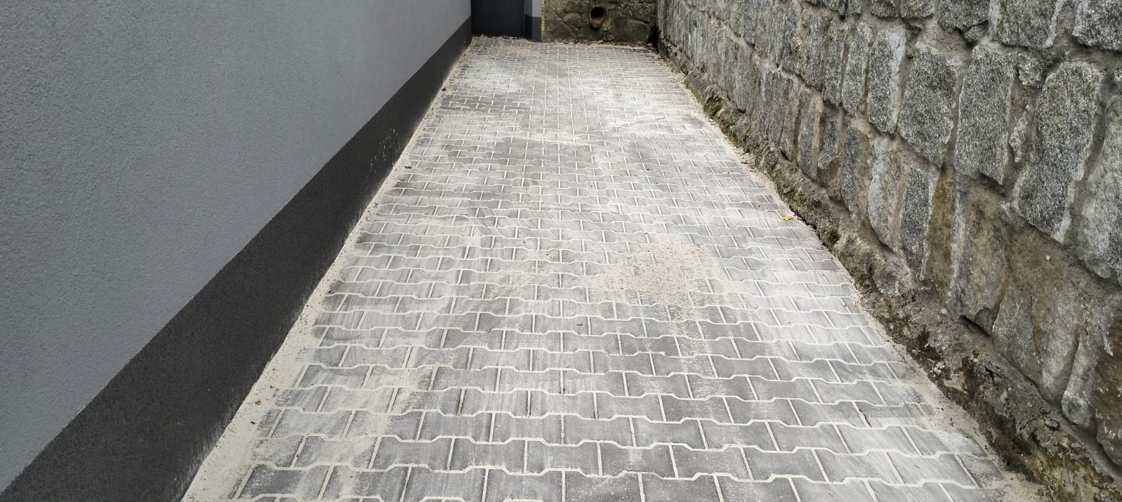 Ułożona szara kostka brukowa typu I na wąskim chodniku między szarą ścianą budynku a kamiennym murem. Widok z góry, lekko z ukosa.