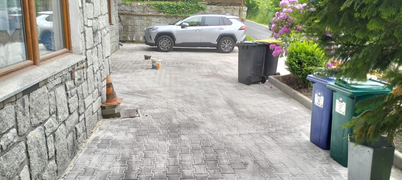 Ułożona szara kostka brukowa na podjeździe, z zaparkowanym srebrnym autem w tle. Widoczne kosze na śmieci i budynek z kamienia.