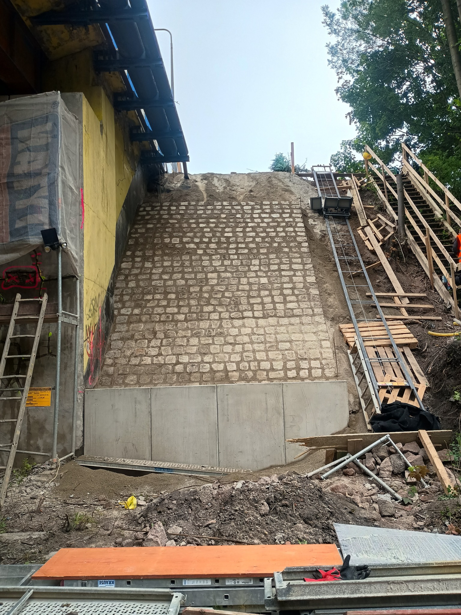 Wzmocnienie skarpy blokami betonowymi i kamienną kostką. Widoczna drabina transportowa i schody. Prace ziemne w toku.