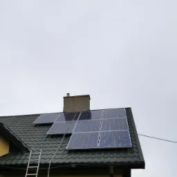 Panele fotowoltaiczne zamontowane na zielonym dachu z blachodachówki, widoczna drabina oparta o ścianę budynku, pochmurne niebo w tle.