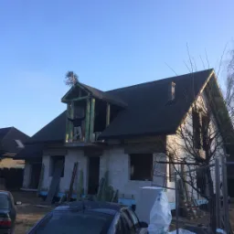 Dom w trakcie budowy z widocznym szkieletem dachu i oknem balkonowym, na balkonie stoi osoba z rozłożonymi rękami, na dachu mała choinka, w tle błękitne niebo i zaparkowany samochód.