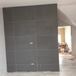 Ściana wyłożona dużymi szarymi płytkami w trakcie remontu łazienki, widoczne puszki elektryczne, podłoga imitująca drewno i materiały budowlane w tle.