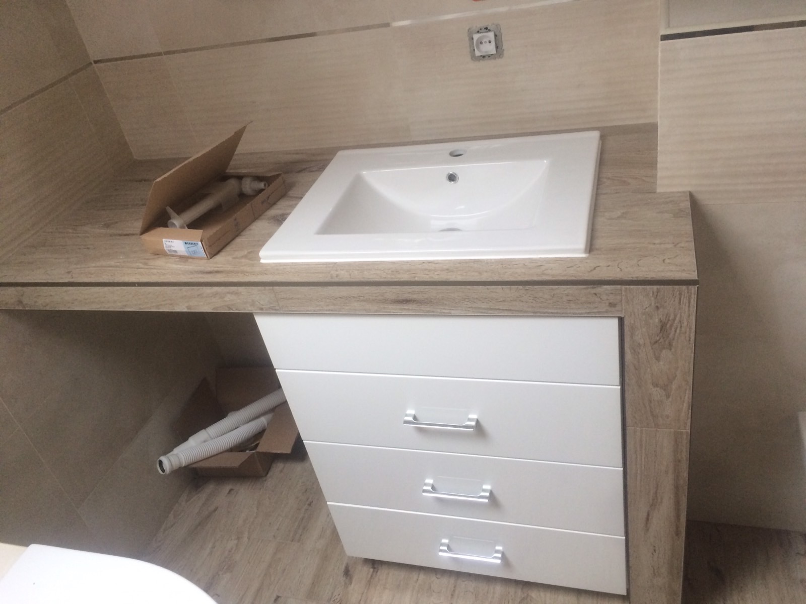 Biała umywalka nablatowa z szafką z czterema szufladami w trakcie montażu. Widoczne akcesoria w kartonach i fragment toalety.