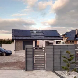 Dom jednorodzinny z zamontowanymi panelami słonecznymi na dachu, widok z zewnątrz, brama wjazdowa, szary samochód osobowy zaparkowany na podjeździe.