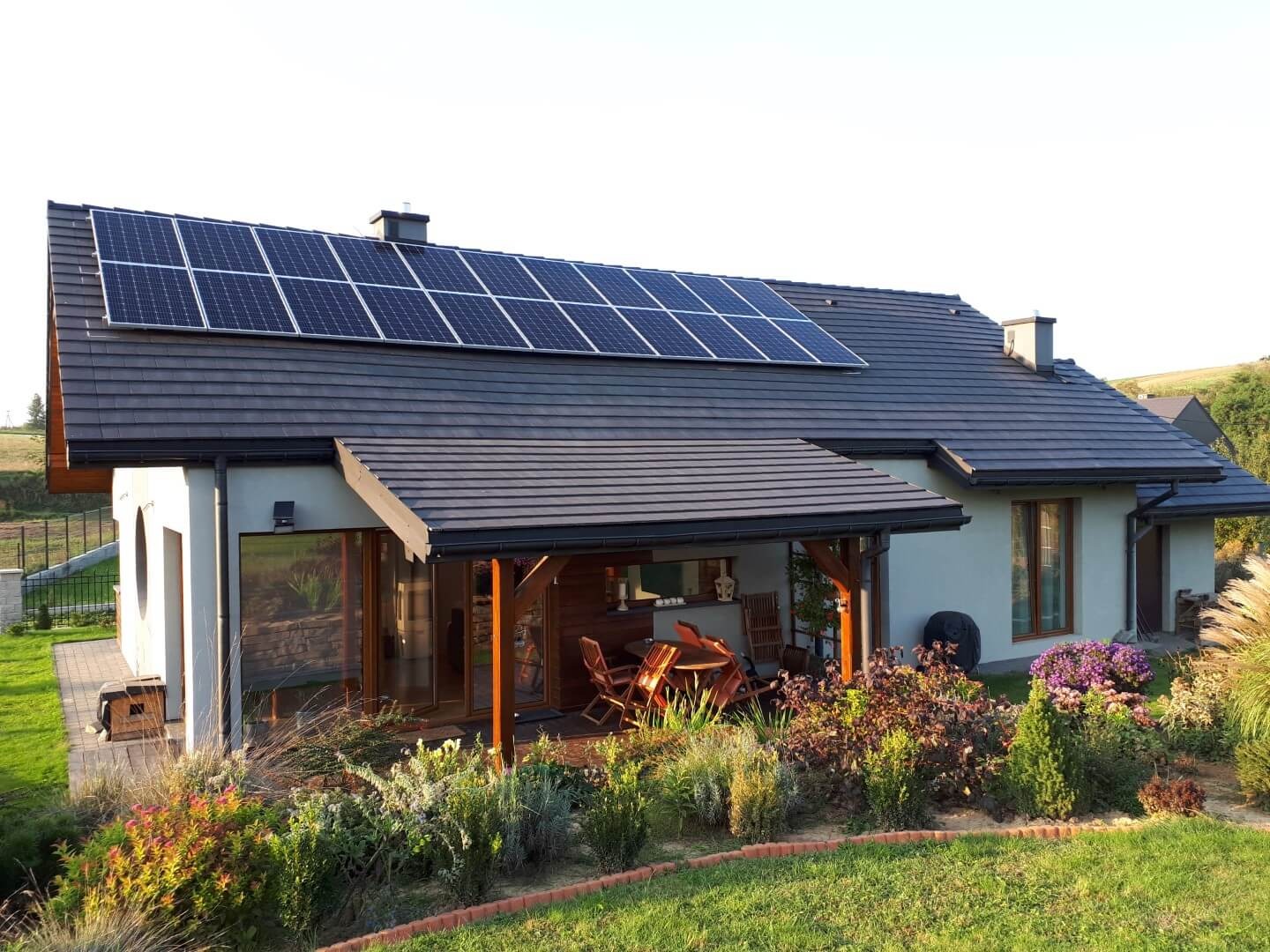 Dom jednorodzinny z zainstalowanymi panelami słonecznymi na dachu, widocznymi z poziomu ogrodu z bujną roślinnością.