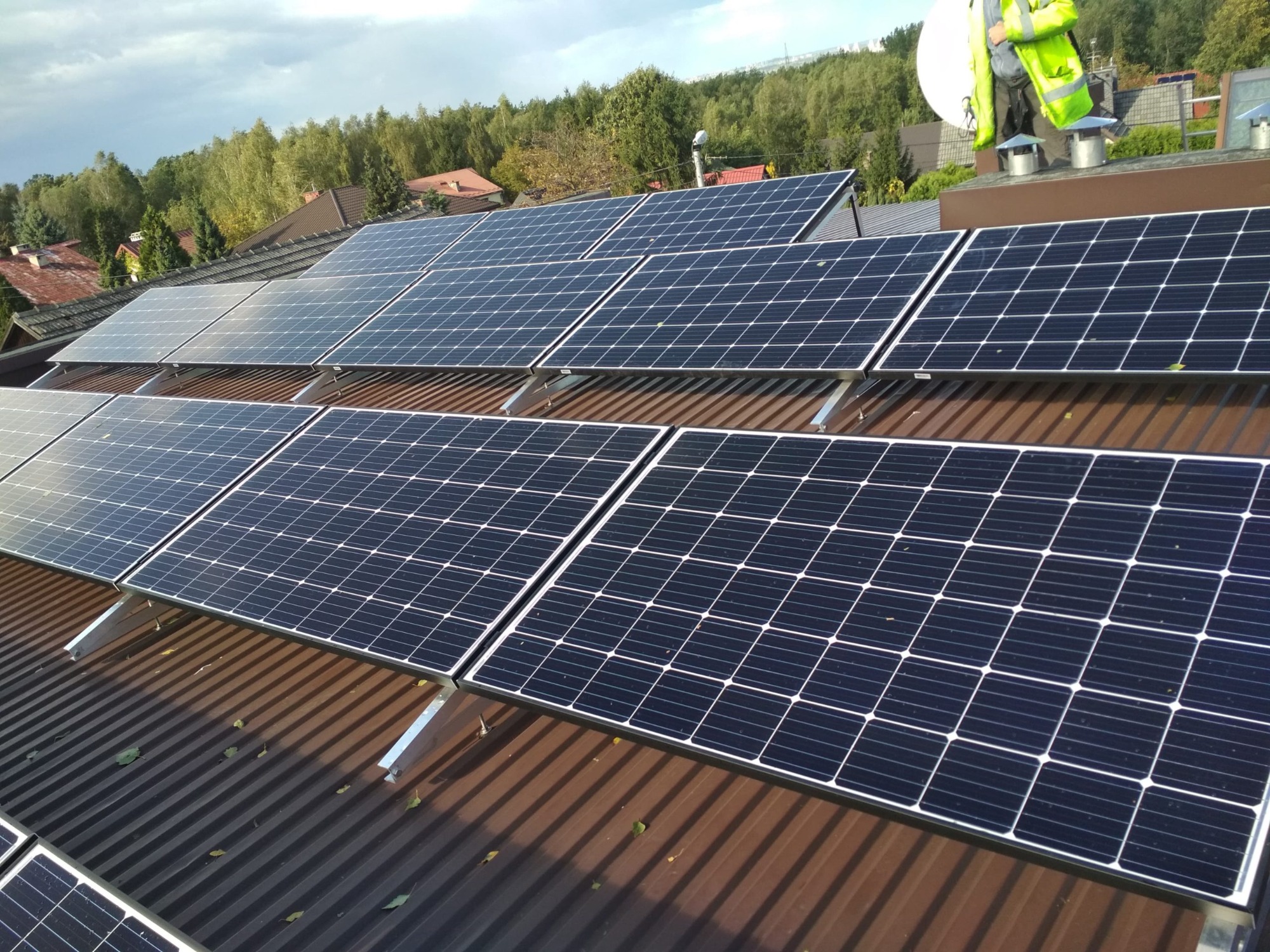 Panele słoneczne na brązowym dachu z blachy trapezowej, widoczny fragment instalatora w jaskrawej kamizelce i antena satelitarna w tle.