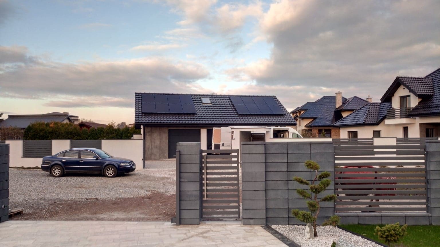 Dom jednorodzinny z zamontowanymi panelami słonecznymi na dachu, widok z zewnątrz, brama wjazdowa, szary samochód osobowy zaparkowany na podjeździe.