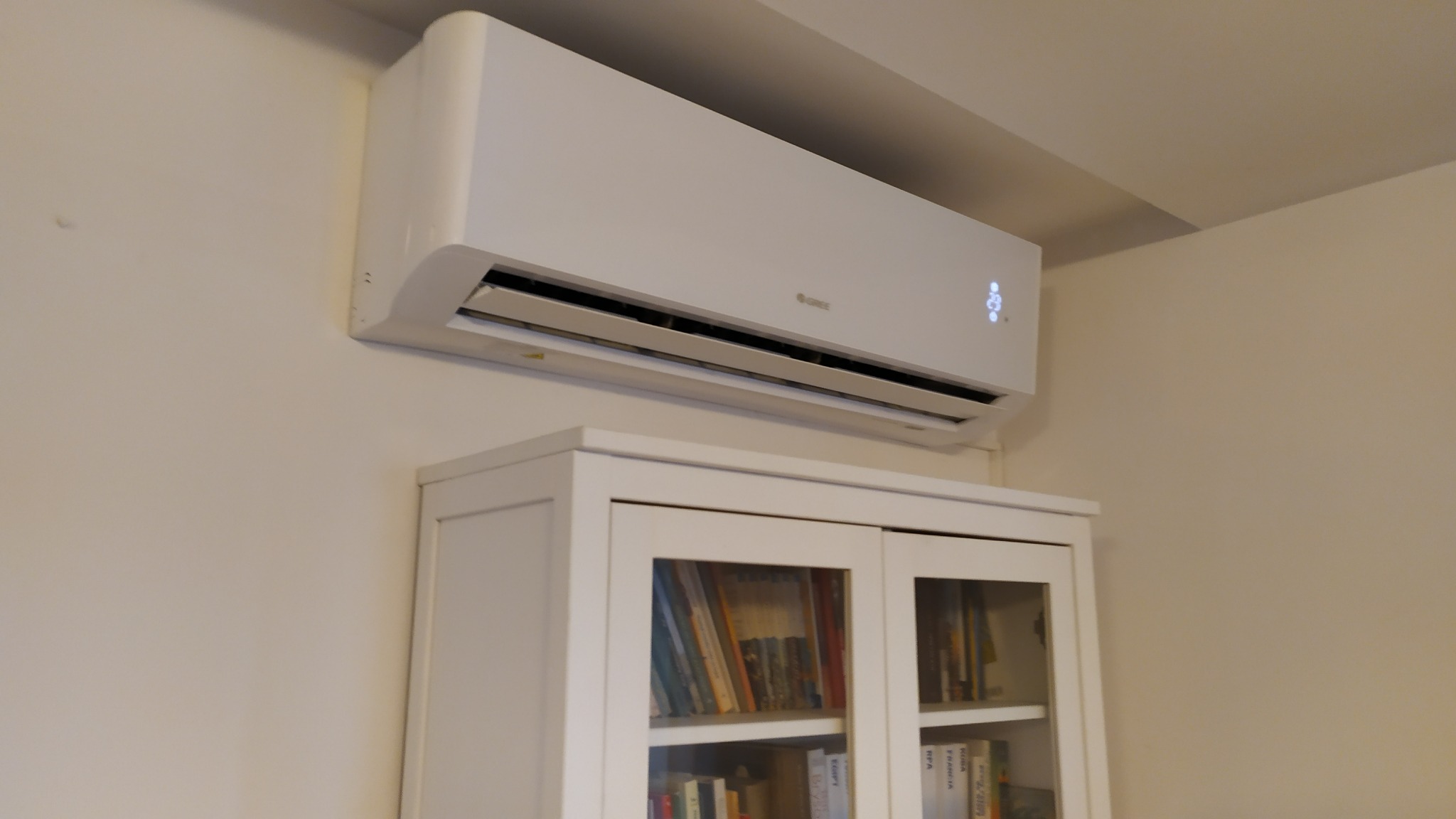 Biały klimatyzator marki Gree zamontowany na ścianie nad białym regałem z książkami, widoczne cyfry na wyświetlaczu urządzenia.