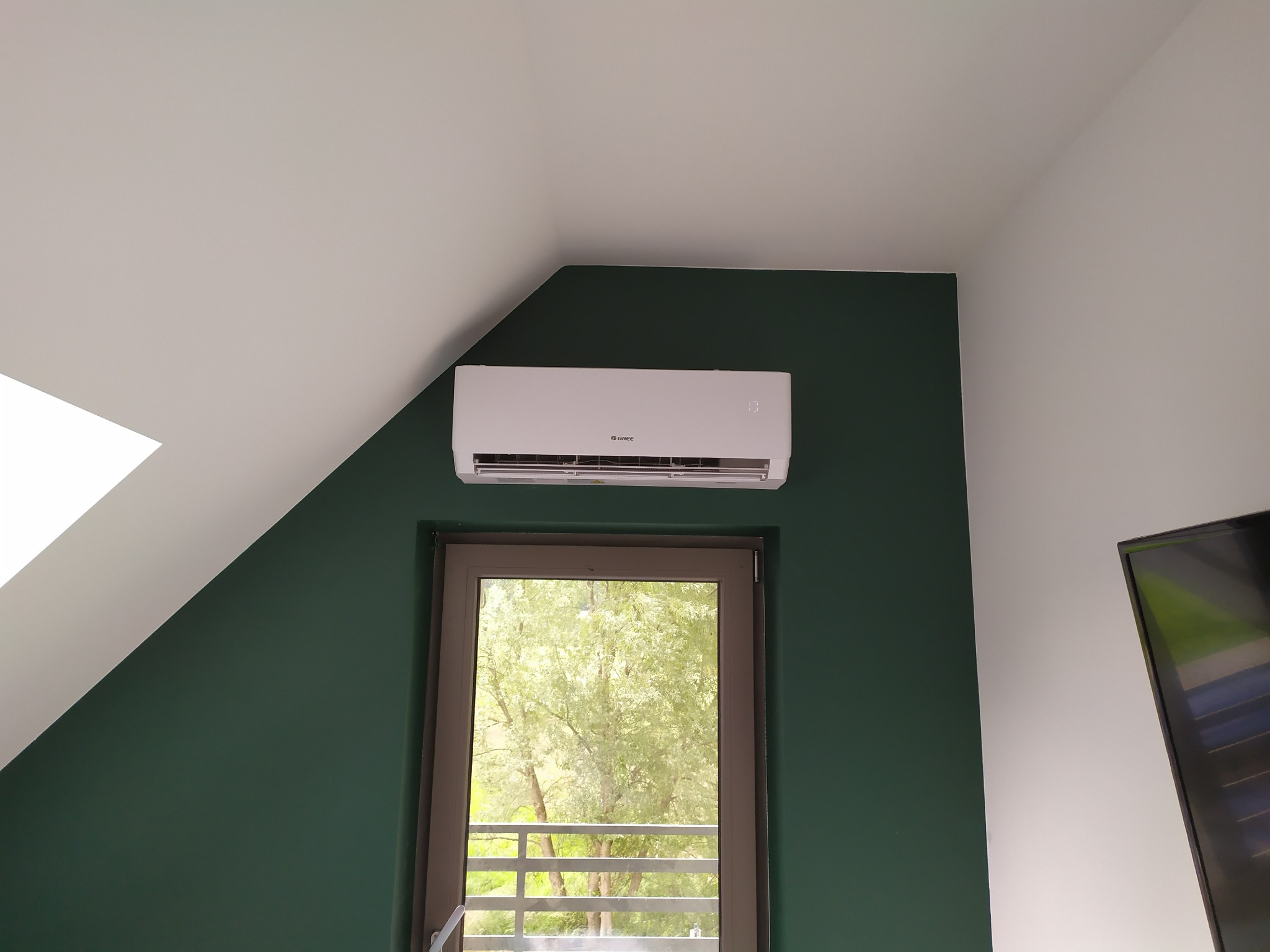 Biały klimatyzator marki Gree zamontowany nad oknem w pokoju z zieloną ścianą i białym skośnym sufitem.