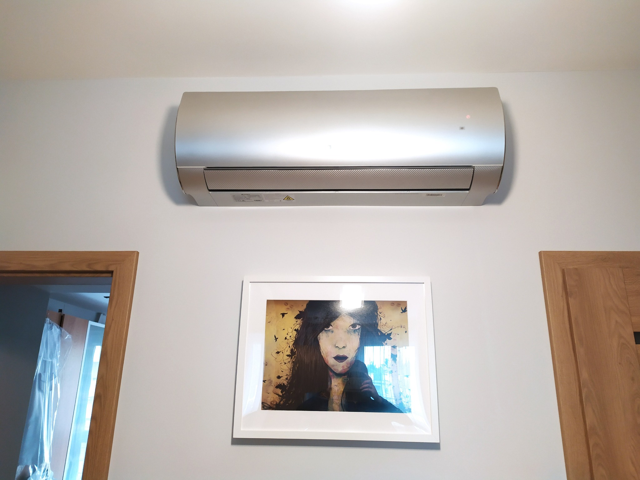 Srebrny klimatyzator ścienny zamontowany nad obrazem w białej ramie, widoczne fragmenty drewnianych ościeżnic drzwi po bokach.