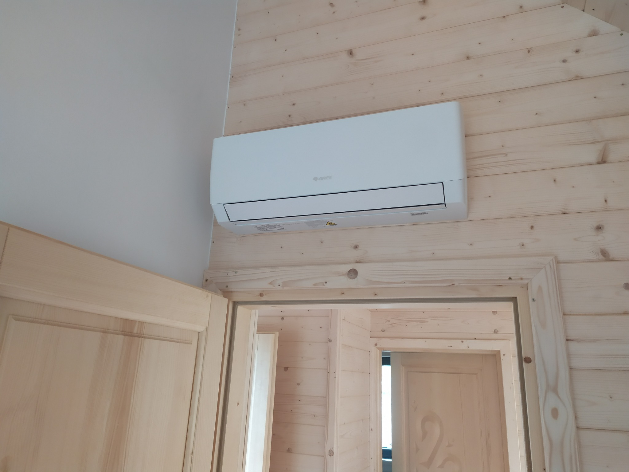 Biały klimatyzator marki Gree zamontowany na ścianie z boazerii w drewnianym domu, widoczny fragment ościeżnicy drzwi.