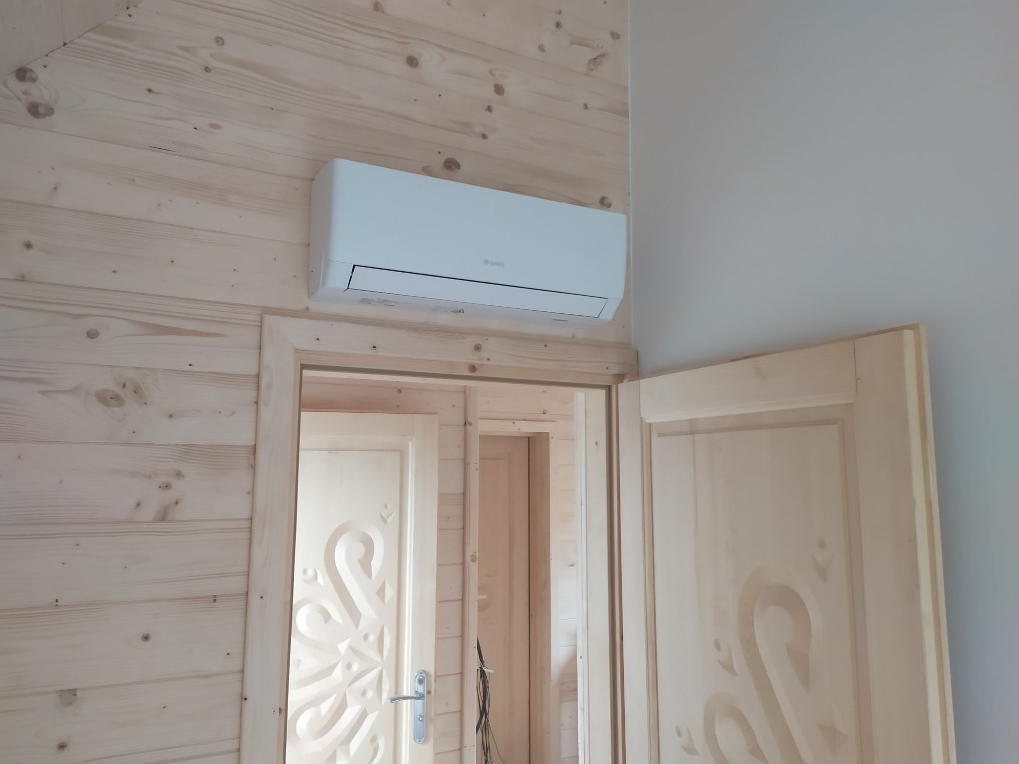 Biały klimatyzator marki Gree zamontowany na drewnianej ścianie z boazerii, widok na otwarte drewniane drzwi z dekoracyjnym wzorem.
