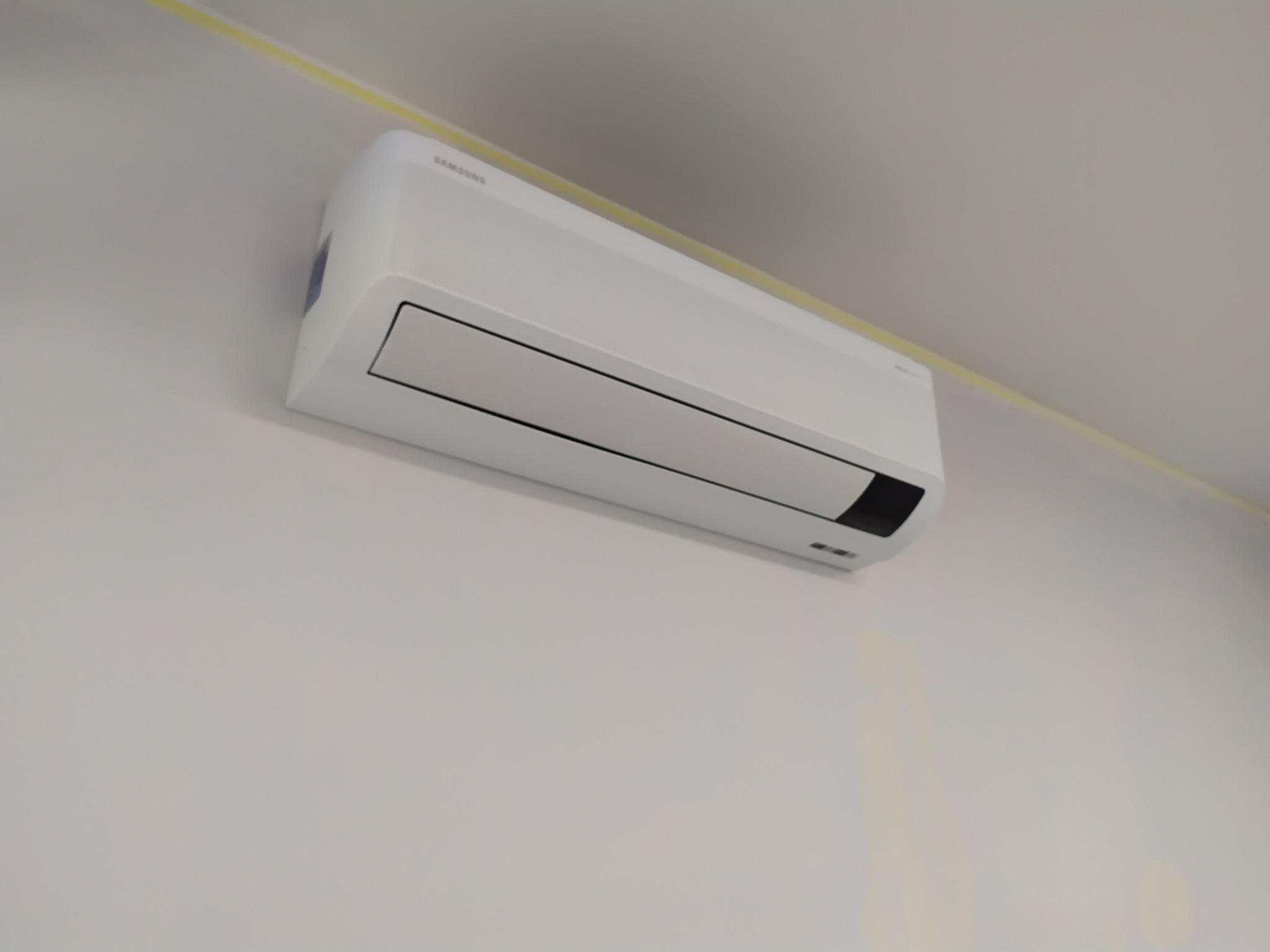 Biała jednostka wewnętrzna klimatyzatora Samsung zamontowana na jasnej ścianie, widoczna listwa sufitowa w kolorze żółtym.
