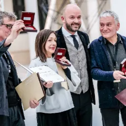 Grupa osób pozuje do zdjęcia, prezentując nagrody w postaci medali w pudełkach i dyplomów podczas uroczystości.