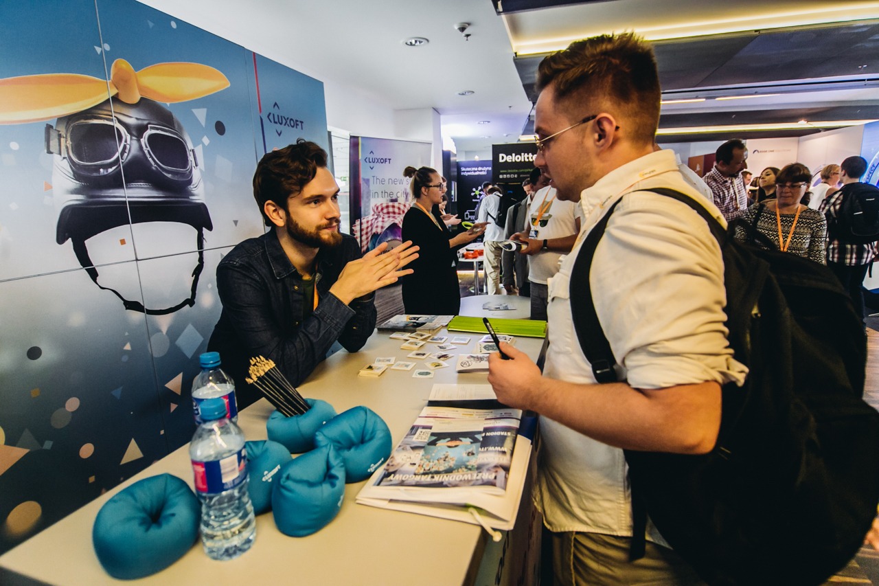 Mężczyzna z brodą rozmawia z klientem przy stoisku Luxoft na evencie, w tle grafika z hełmem lotniczym.