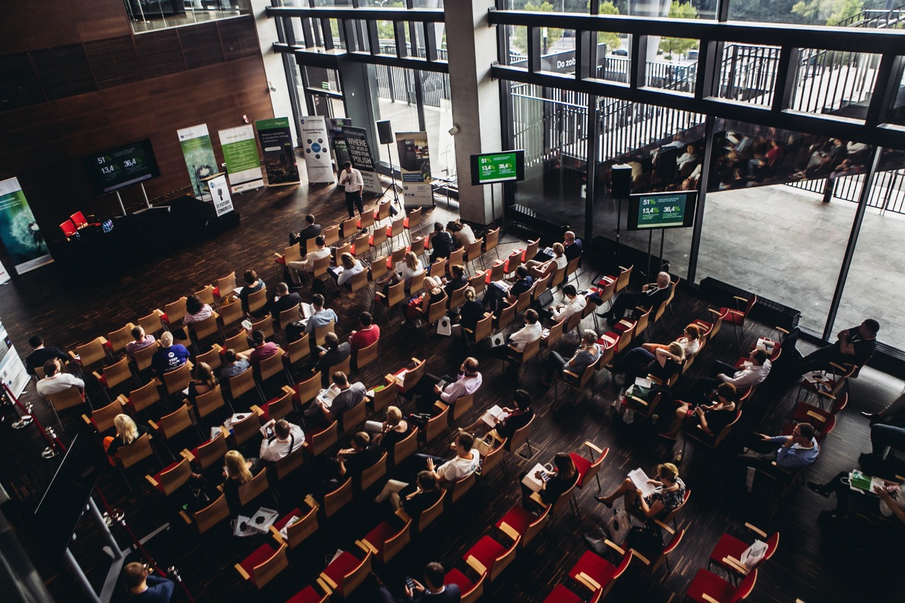 Widok z góry na salę konferencyjną z uczestnikami siedzącymi na krzesłach i prelegentem na scenie, widoczne ekrany z danymi statystycznymi i roll-upy reklamowe w tle.