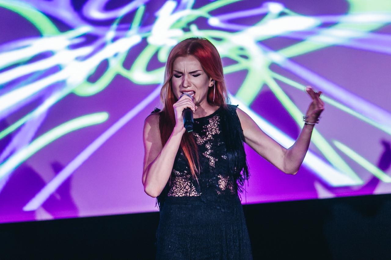 Piosenkarka z rudymi włosami śpiewa do mikrofonu na scenie z fioletowym, abstrakcyjnym tłem.