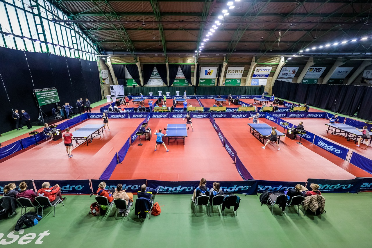 Widok z góry na halę sportową z kilkoma stołami do tenisa stołowego, na których rozgrywane są mecze, z widownią siedzącą wzdłuż boiska, w tle widoczne banery reklamowe.