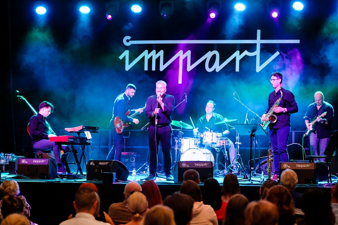 Zespół muzyczny grający na scenie podczas koncertu, oświetlony niebieskim i fioletowym światłem, widoczna publiczność.