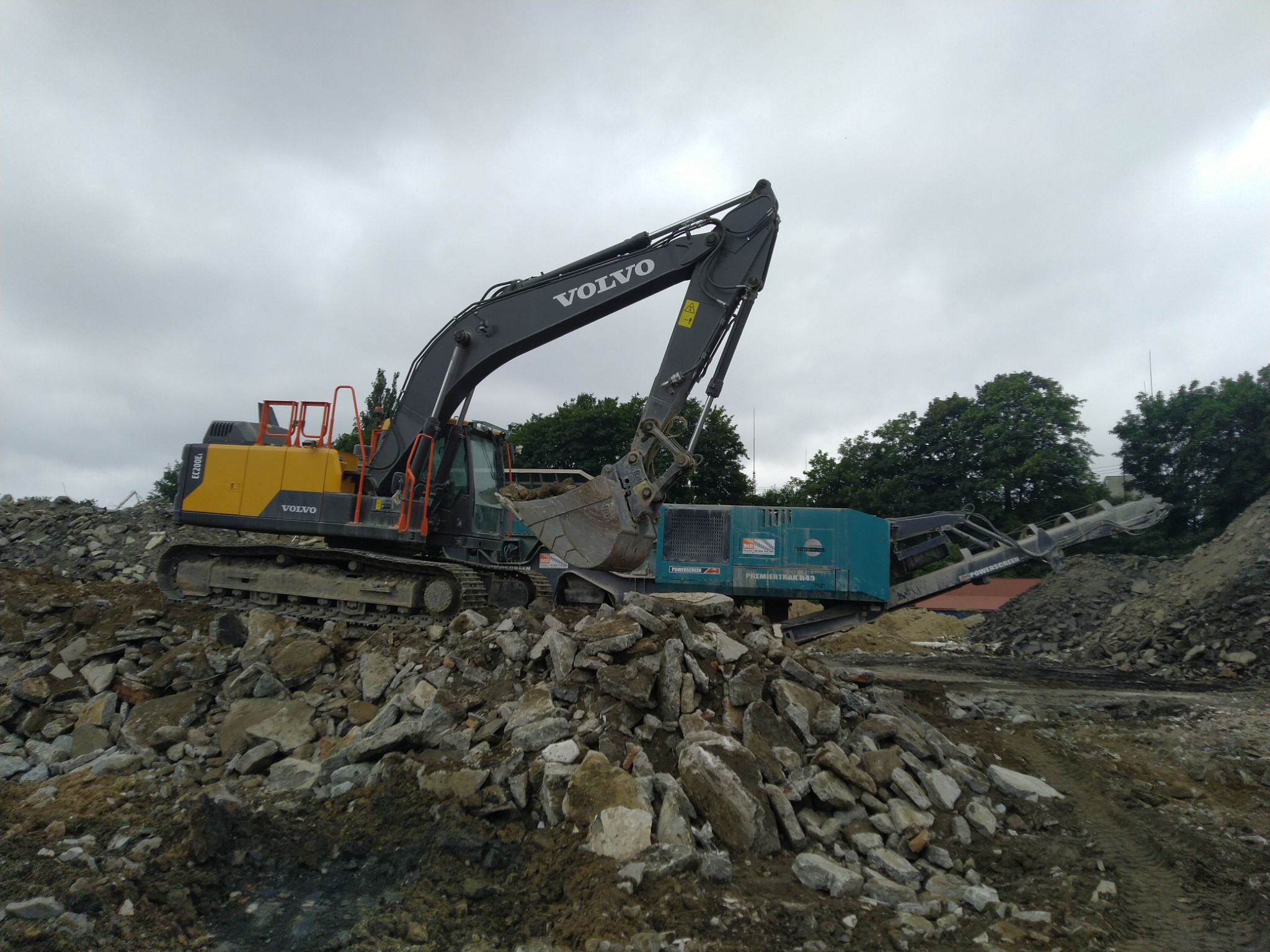 Żółto-szara koparka Volvo na szczycie sterty gruzu i ziemi, obok przesiewacz do kruszyw Premier Track 849, pochmurne niebo w tle.