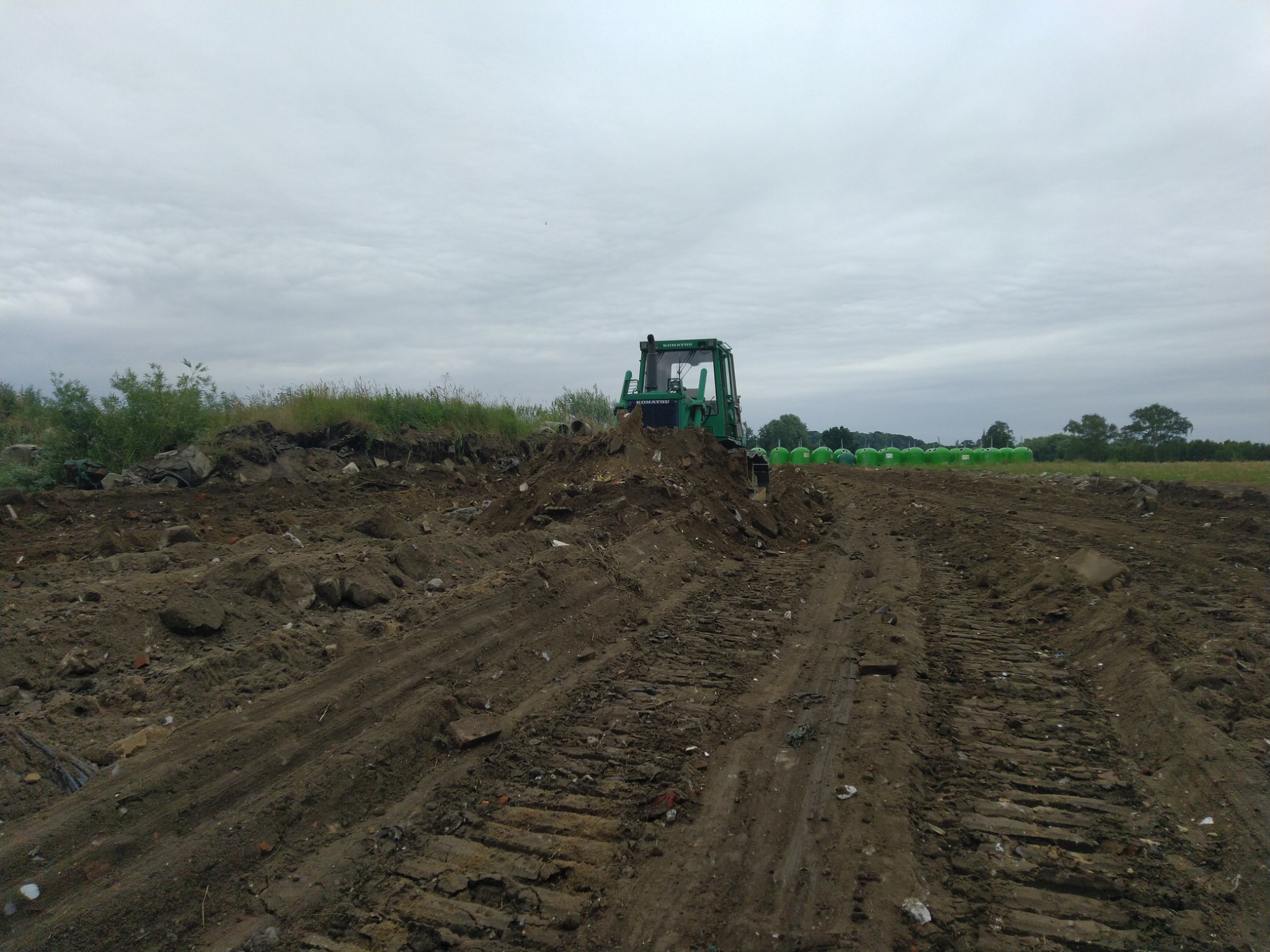 Zielony traktor Komatsu na nierównym, brązowym terenie z widocznymi śladami gąsienic, w tle rząd zielonych zbiorników i pochmurne niebo.