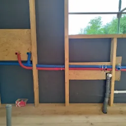 Wewnętrzna instalacja wodna w trakcie montażu: rury czerwone i niebieskie przymocowane do drewnianej konstrukcji ściany, widoczne elementy kanalizacji i pudełko z akcesoriami.