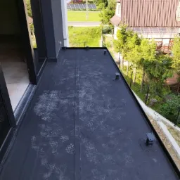 Czarna, płaska powierzchnia dachu balkonu z widocznymi śladami butów, zabezpieczona czarną blachą, z elementami mocującymi wystającymi ku górze. W tle widok na domy i zieleń.