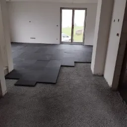 Wnętrze w trakcie prac: rozłożone czarne maty izolacyjne na podłożu z szarego styropianu pod ogrzewanie podłogowe, widok na okno z zewnątrz.