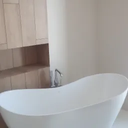 Wolnostojąca, biała wanna o owalnym kształcie z chromowaną baterią, umieszczona w minimalistycznej łazience z drewnianą zabudową.