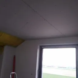 Montaż płyt kartonowo-gipsowych na suficie w pokoju z oknem, widoczna drabina i izolacja z wełny mineralnej na ścianie, surowy stan wykończenia.