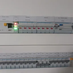 Dwa rzędy bezpieczników w skrzynce rozdzielczej, górny rząd marki Eaton z trzema świecącymi diodami, dolny rząd marki Hager z opisami obwodów.