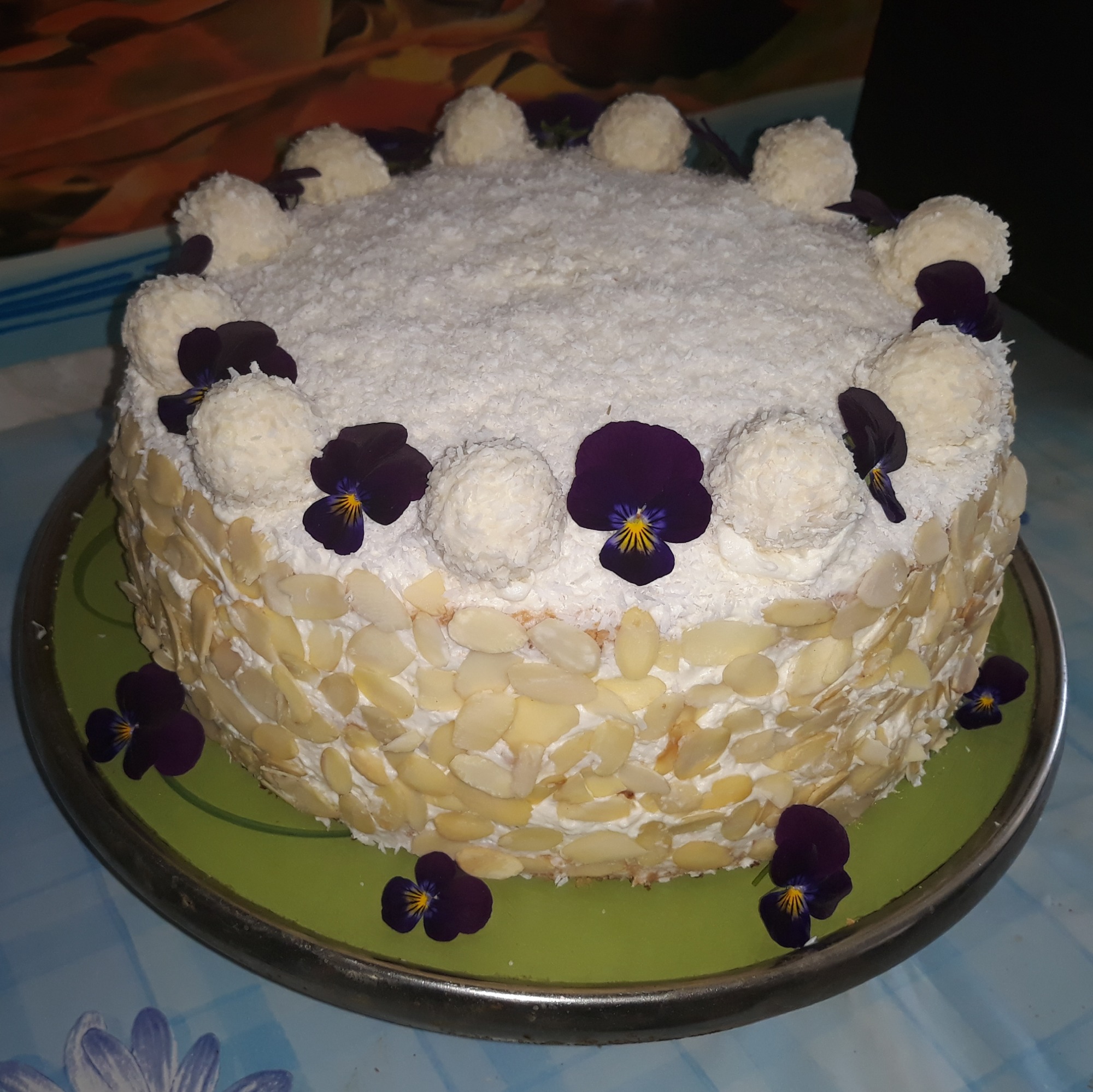 Okrągły tort kokosowy z płatkami migdałów i dekoracją z fiołków na zielonym talerzu.