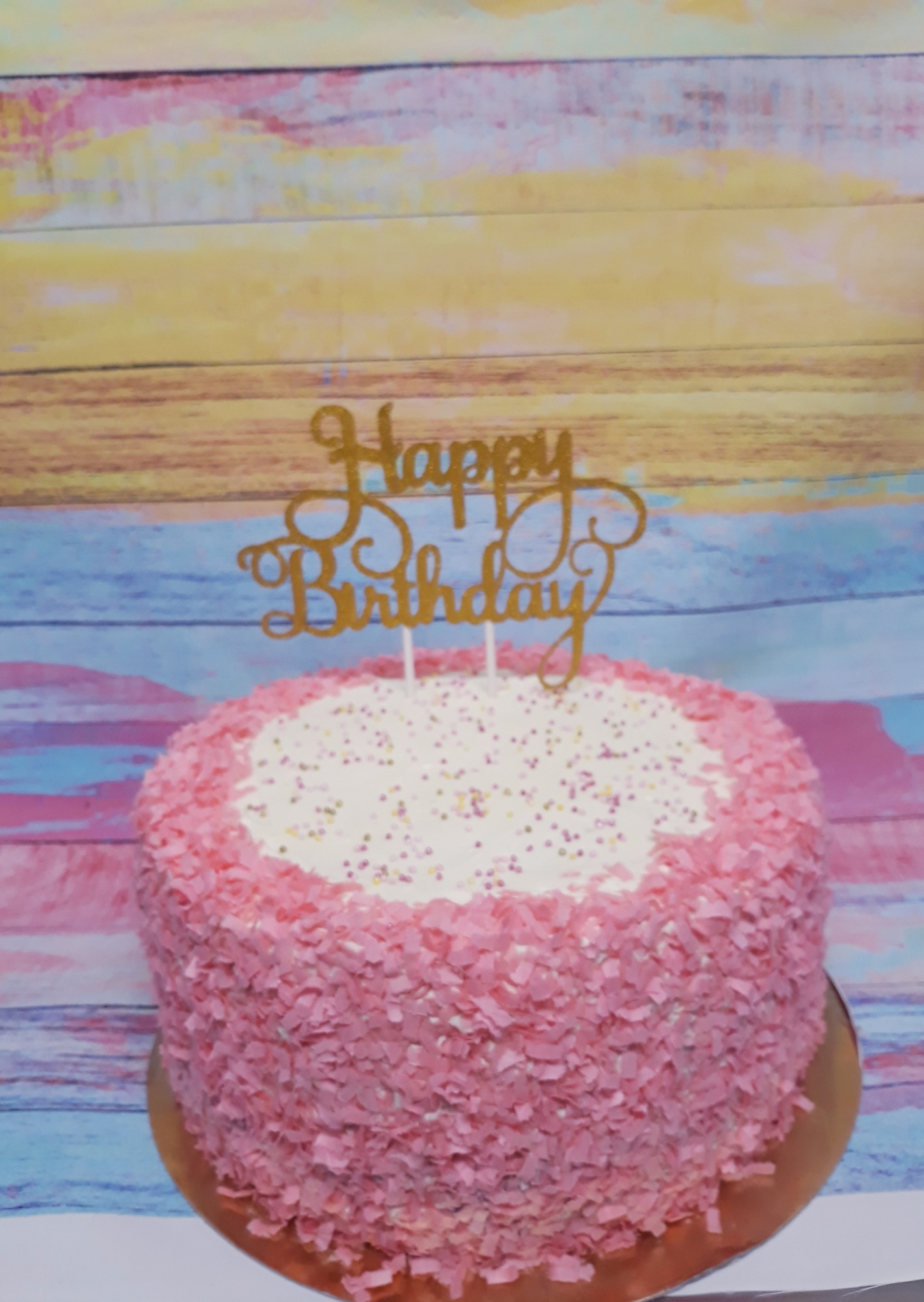 Tort urodzinowy z różową posypką kokosową i złotym napisem 'Happy Birthday' na tle kolorowej drewnianej ściany.