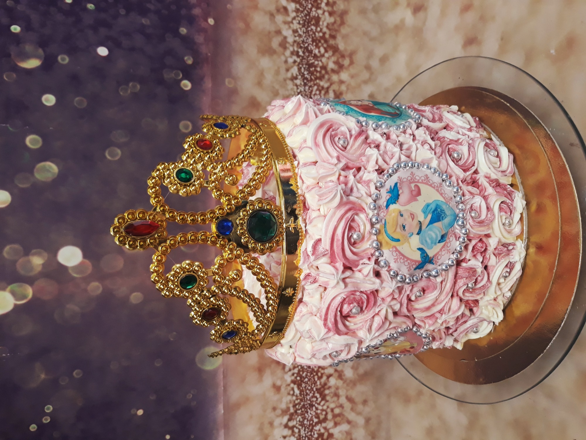 Tort urodzinowy udekorowany kremem w kształcie róż, z jadalną koroną wysadzaną kolorowymi kamieniami i wizerunkami postaci z bajek, umieszczony na szklanej paterze.