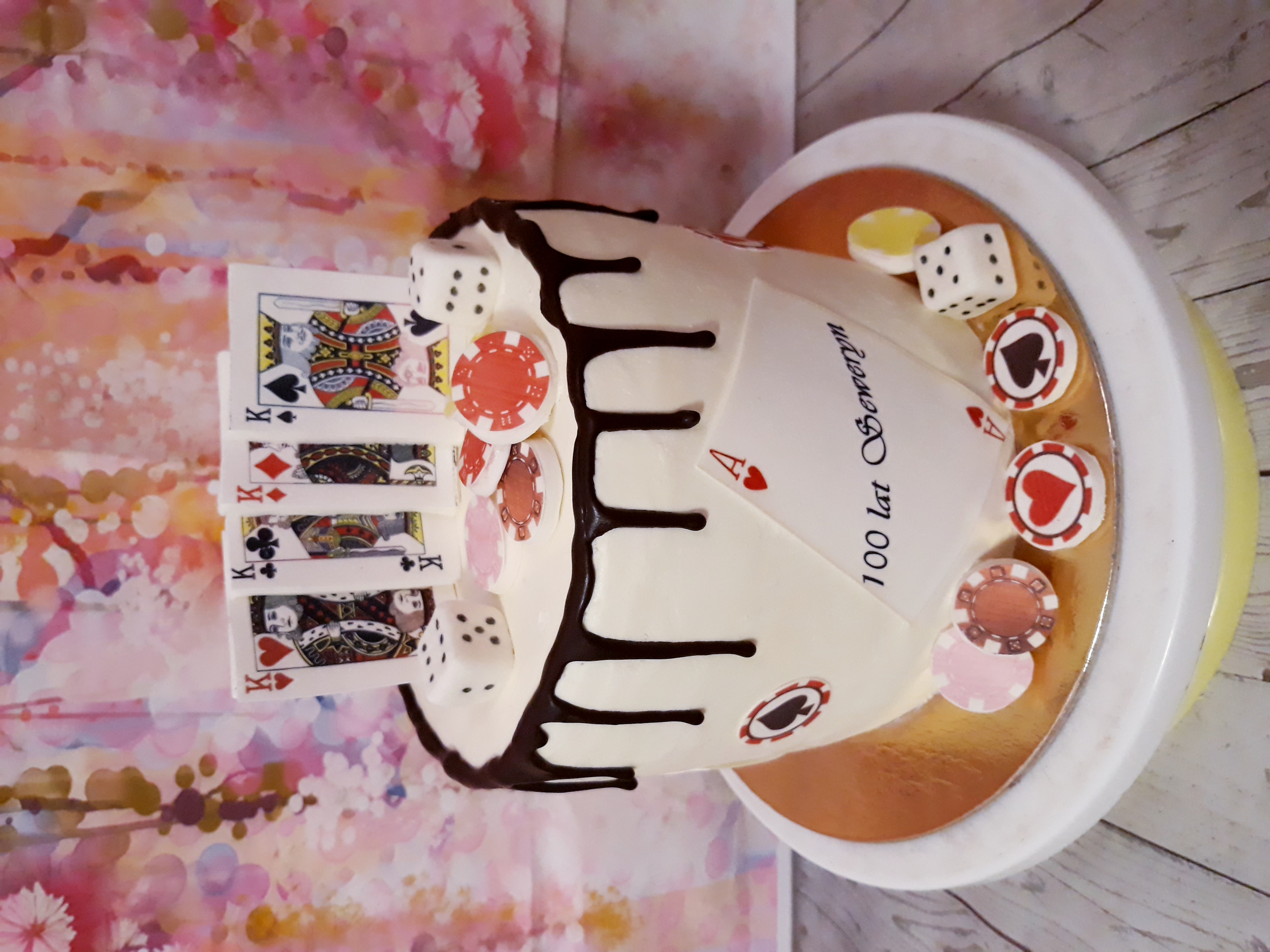 Tort urodzinowy w stylu kasyna z kartami Królów, kostkami, żetonami i polewą czekoladową na tle abstrakcyjnym.