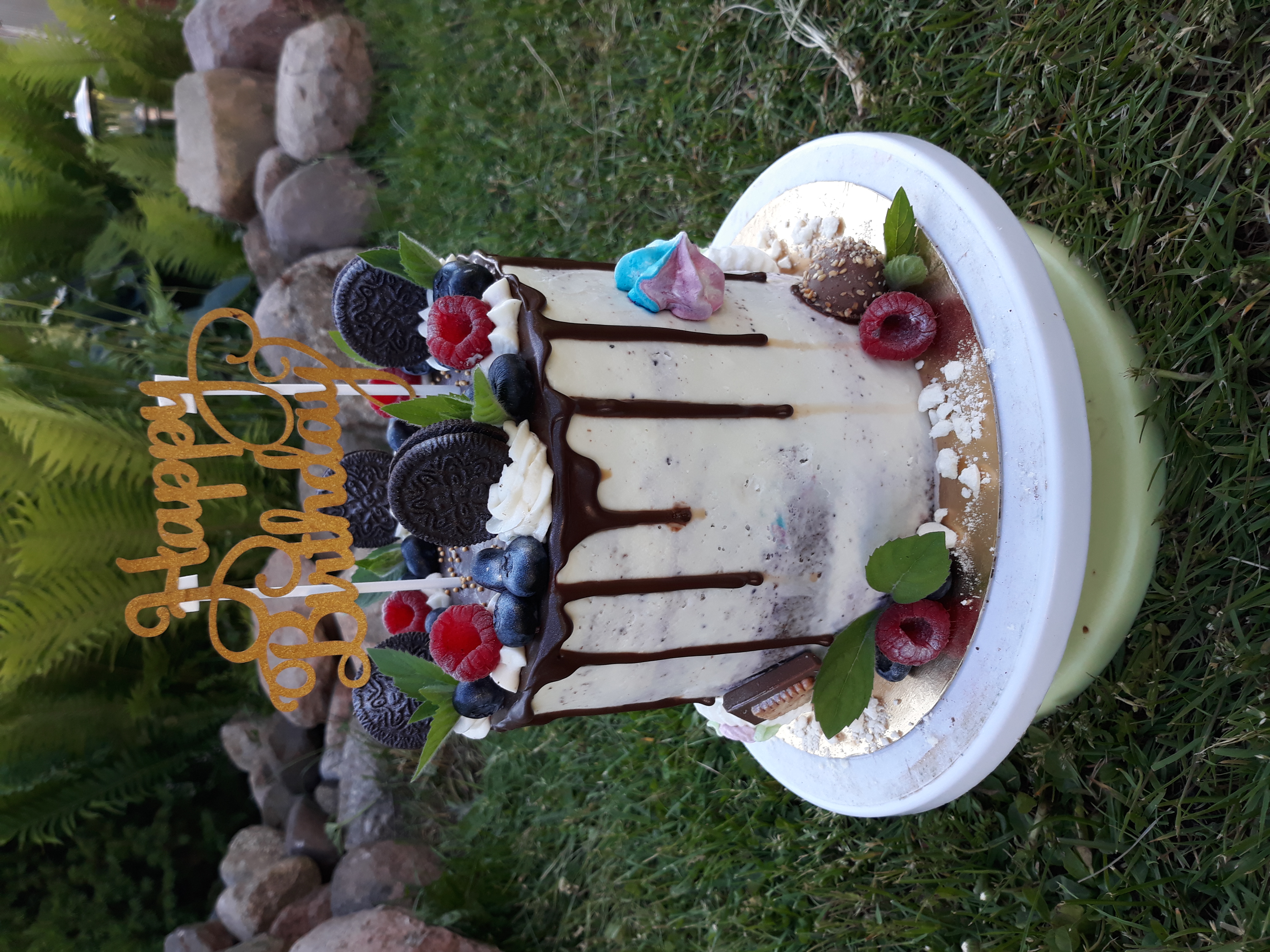 Tort urodzinowy udekorowany czekoladą, borówkami, malinami, ciasteczkami Oreo i napisem 'Happy Birthday' na tle trawy i kamieni.
