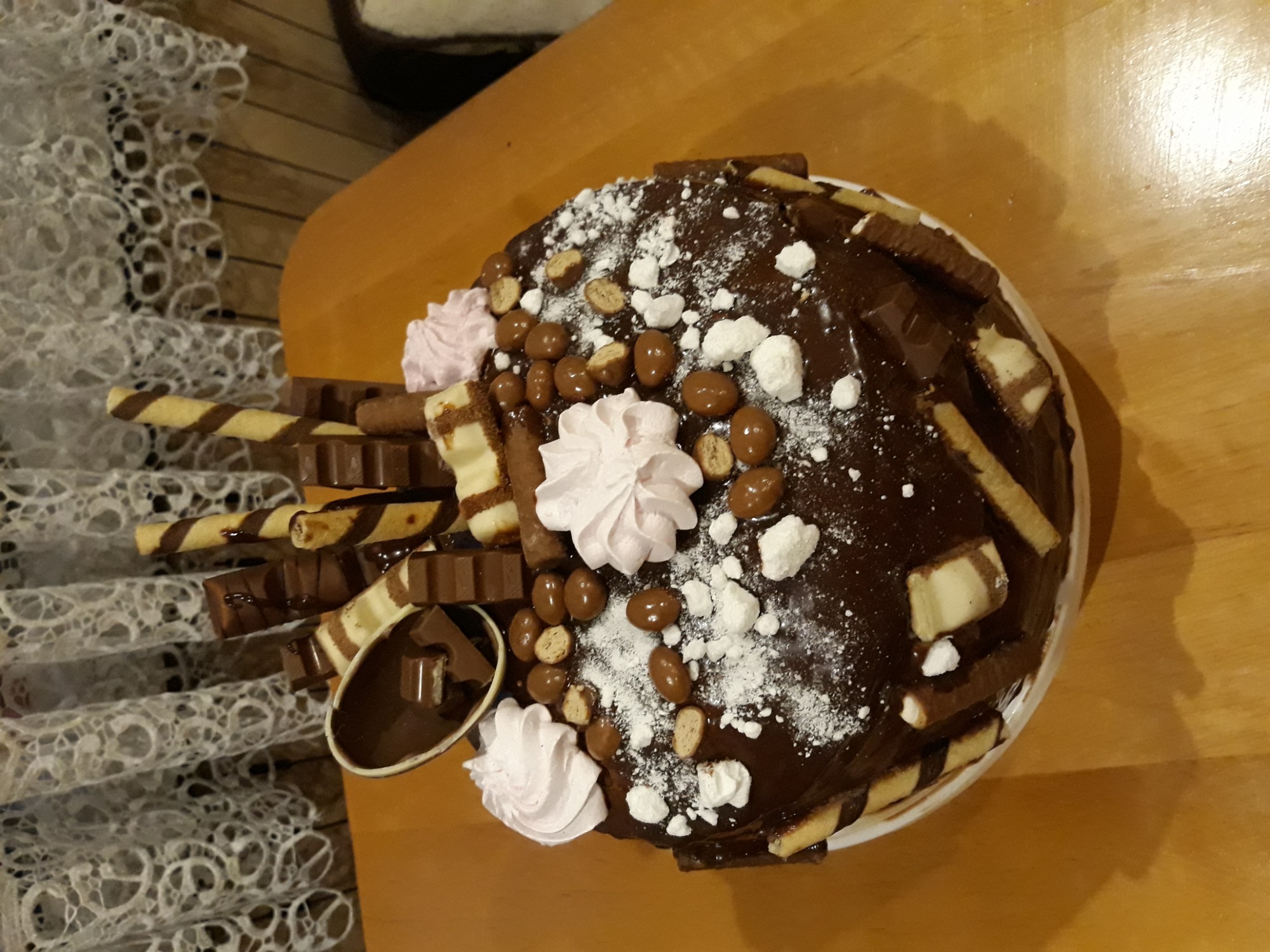 Tort czekoladowy z batonikami, waflami, kremem i posypką, częściowo pokrojony, na drewnianym stole z koronkową serwetą.