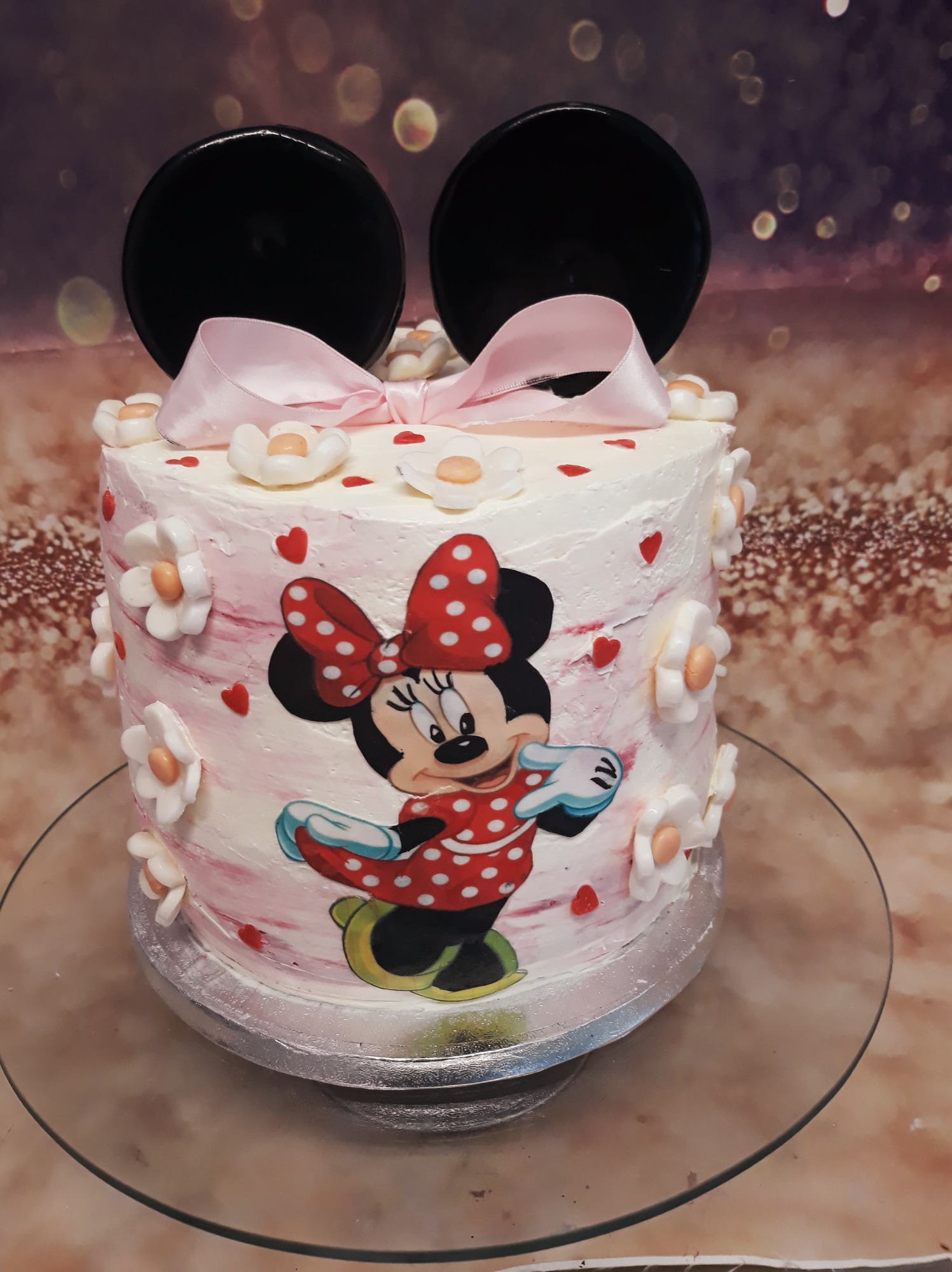 Tort udekorowany motywem Myszki Minnie, z białym kremem, różowymi akcentami, małymi kwiatkami i czarnymi uszami z opłatka, na szklanym talerzu.